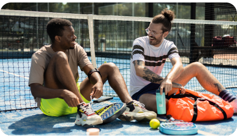 Photo of two men sat in a Padel Tennis court, laughing after playing a game. 